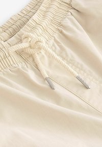 Shorts beige clair avec une taille élastique, détail de nœud de cordon de serrage, et accents en métal. Tissu lisse avec un léger éclat et des détails de couture.