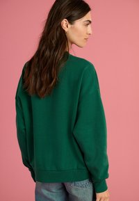 Zelena dukserica s opuštenim krojem i rebrastim manžetama, koja ima okrugli izrez i izrađena je od mekog pamučnog materijala. Model prikazan sa stražnje strane.