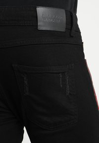 Jean de mezclilla negra con detalles desgastados, que cuentan con una etiqueta de cuero que dice "GLORIOUS GANGSTA" en la cinturilla y acentos rojos en los laterales.