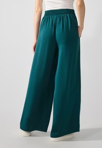 Pantalons à jambes larges en teal, avec une taille élastique, une texture lisse et des poches latérales. Le tissu présente un motif de froissé subtil.