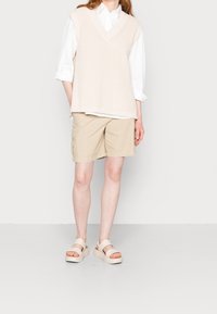 Persona che indossa un maglioncino senza maniche beige in maglia sopra una camicia bianca a maniche lunghe, pantaloni cargo color beige e sandali con plateau beige, in piedi su uno sfondo bianco.
