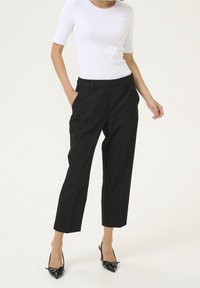 Pantalon noir sur mesure avec plis, poches latérales et coupe fuselée, accompagné d'un haut blanc ajusté à manches courtes et de chaussures noires à talons.