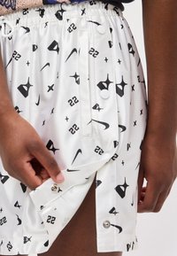 Κοντινό πλάνο μαύρων χεριών που προσαρμόζουν κουμπιά τύπου snap σε άσπρες βερμούδες με μαύρα λογότυπα Nike και τον αριθμό 22.