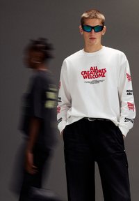 Weißes Langarmhemd mit rotem Text "ALLE KREATUREN WILLKOMMEN" und Grafiken auf den Ärmeln; kombiniert mit schwarzen Cargo-Hosen; der Hintergrund zeigt eine verschwommene Figur.