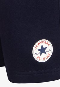 Pantaloni corti in cotone blu navy con una toppa circolare bianca, decorata con una stella blu e testo rosso, che riporta "CONVERSE ALL STAR" e "Chuck Taylor."
