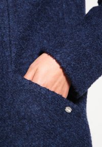 Tessuto di un cappotto blu con texture, caratterizzato da una tasca laterale, con un dettaglio di bottone metallico lucido e una superficie morbida e pelosa.