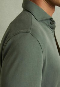 Cămașă scurtă cu mâneci, de culoare verde închis, cu guler, având un închizător cu fermoar și un material neted, texturat. Detalii vizibile ale cusăturii pe umăr.