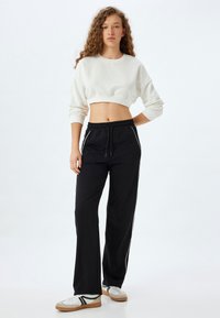 Felpa bianca corta con maniche lunghe, abbinata a pantaloni neri a gamba larga con strisce bianche laterali e vita elastica. Sneakers casual incluse.