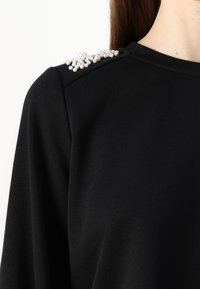 Μαύρη μπλούζα με λεία υφή, που διακοσμείται με μια συστάδα λευκών στρας στον αριστερό ώμο.