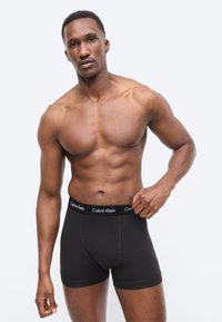 Muskulárny muž v čiernych boxerkách Calvin Klein stojaci pred jednoduchým svetlým pozadím, pravou rukou si upravuje pás.