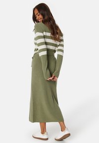 Abito a maglia verde a righe con maniche lunghe e vita regolabile, caratterizzato da ampie righe bianche e un tessuto texturizzato. Completato con sneakers bianche.
