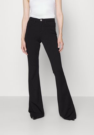 Vrouw die zwarte, hoge taille, uitlopende broek draagt met knoop en rits, gecombineerd met een wit mouwloos topje tegen een effen achtergrond.