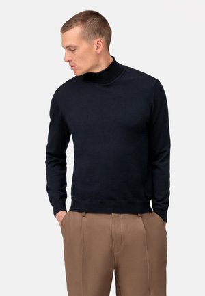 Marineblauer Rollkragenpullover aus weichem Material, mit langen Ärmeln, einer figurschmeichelnden Passform sowie gerippten Bündchen und Saum, kombiniert mit hellbraunen Hosen.