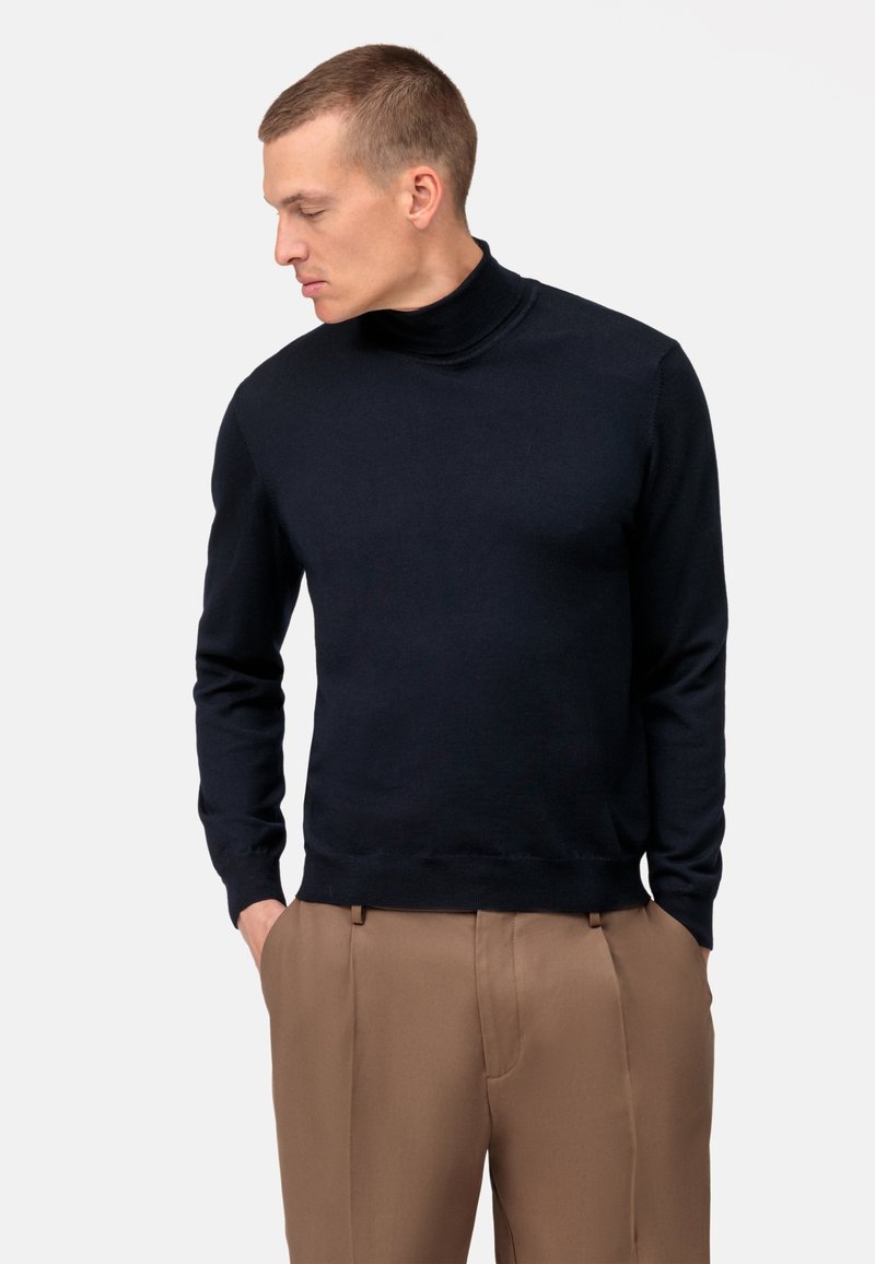 Maglione a collo alto navy in tessuto morbido, con maniche lunghe, una vestibilità aderente e polsini e orlo a coste, abbinato a pantaloni marrone chiaro.
