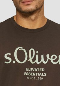 Brown cotton T-shirt featuring a large light green "s.Oliver" logo and "Elevated Essentials Since 1969" text printed in white.