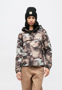 Kaplaarzen anorak jas met een bruin, grijs en beige abstract patroon, voorzien van een voorzak en verstelbare manchetten. Draagt een zwarte beanie.