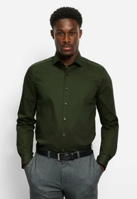 Donkergroene blouse met lange mouwen, klassieke kraag, knopen aan de voorkant en een gladde textuur, gecombineerd met grijze broek en een zwarte riem.