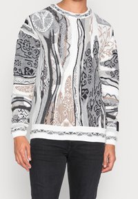 Pull en tricot multicolore avec des motifs complexes gris, blanc et marron, des poignets et un ourlet côtelés, un col rond et une finition texturée.