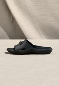 Sandale noire à enfiler avec une semelle intérieure texturée surélevée et un emblème décoratif sur la bride, posée sur une surface beige clair avec des ombres douces.