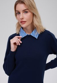 Pull marin à manches longues, avec un col rayé bleu. Gros plan sur une main ajustant le col, montrant une texture lisse et une coupe ajustée.
