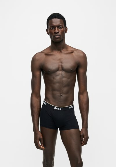 Homme à la peau foncée en forme, debout face à l'avant, portant un boxer noir de la marque BOSS sur un fond blanc uni.