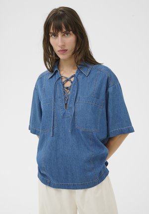 Kvinde iført en blå denimskjorte med korte ærmer, snørelukning foran og to brystlommer, kombineret med lyse bukser, stående mod en hvid baggrund.