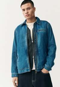 Chaqueta de mezclilla de azul medio con cremallera frontal, dos bolsillos laterales y un bolsillo en el pecho. Presenta un cuello clásico y un corte relajado.