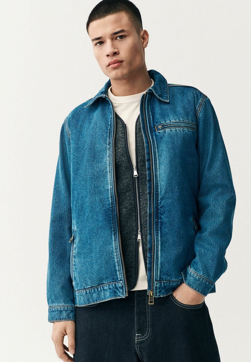 Chaqueta de mezclilla de azul medio con cremallera frontal, dos bolsillos laterales y un bolsillo en el pecho. Presenta un cuello clásico y un corte relajado.