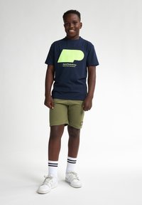 T-shirt bleu marine avec un grand logo lime, associé à un short vert olive. Complété par des baskets blanches et des chaussettes de ville rayées.