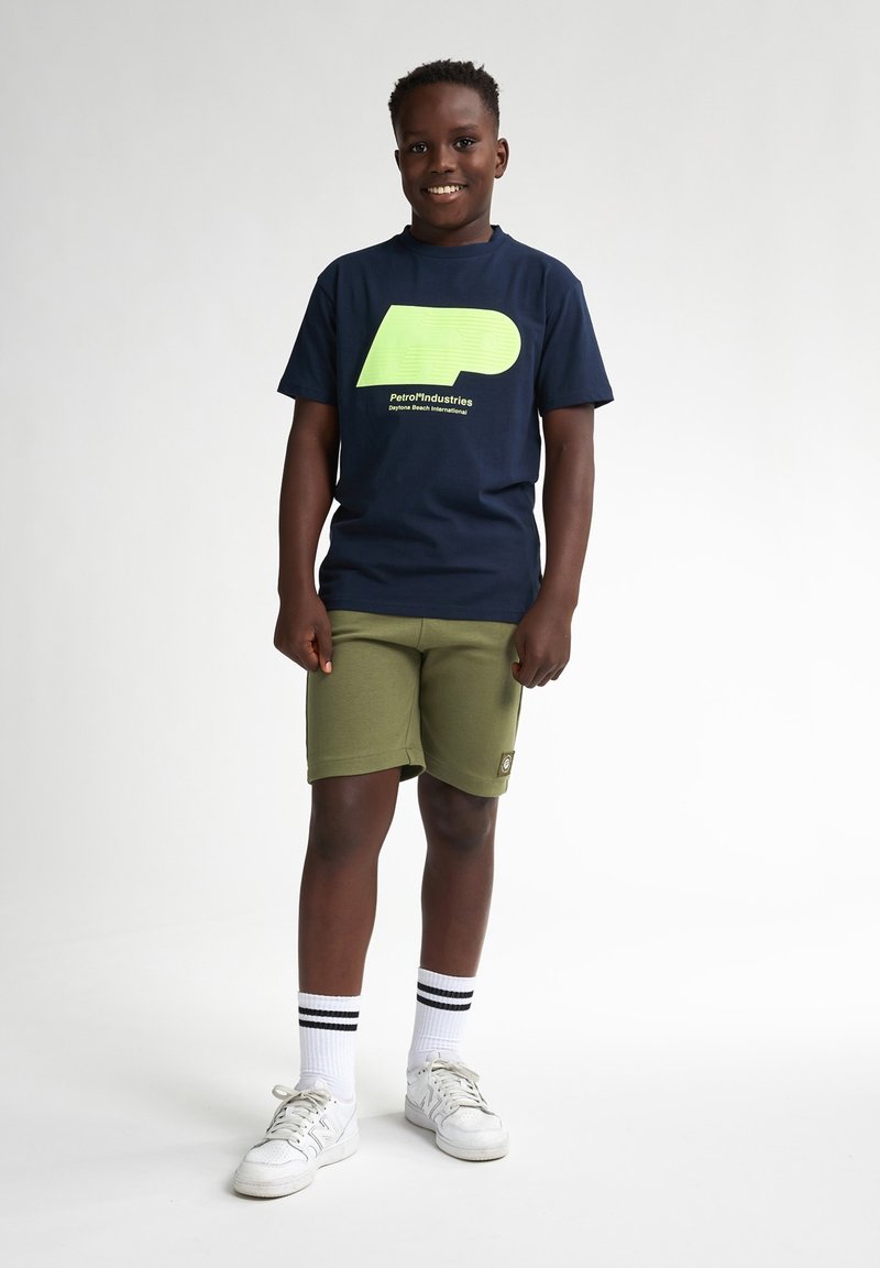 T-shirt bleu marine avec un grand logo lime, associé à un short vert olive. Complété par des baskets blanches et des chaussettes de ville rayées.