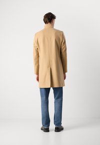 Palton din amestec de lână bej cu guler înalt, ventrață dublă la spate și croială ajustată, purtat peste blugi albaștri și pantofi negri. Design minimalist.