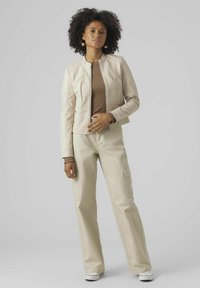 Lichtbeige leren jacket met een korte snit en ritssluiting, gecombineerd met lichtbeige cargobroek met zijzakken en een getextureerd bruin topje.