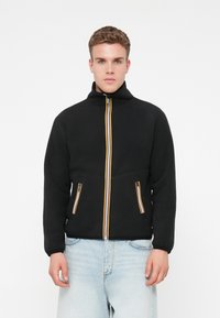 Giovane in giacca di pile nera con zip a strisce e jeans azzurri, in piedi davanti a uno sfondo bianco uniforme.