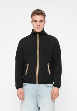 Joven vestido con una chaqueta de forro polar negra con cremalleras a rayas y jeans de color azul claro, de pie contra un fondo blanco liso.