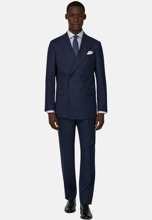 Muž ve tmavě modrém dvouřadém obleku se stejnými kalhotami, bílou košilí, modrou kravatou, bílým kapesníčkem a černými botami.