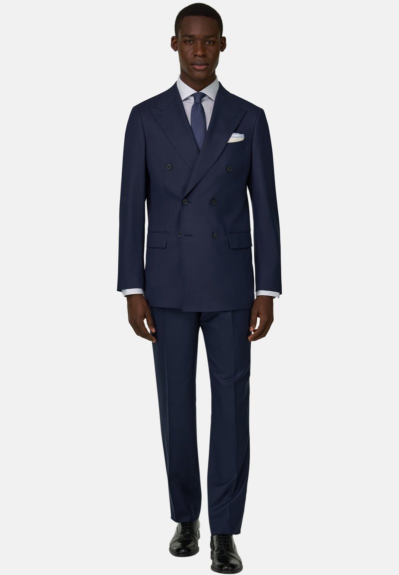 Mann trägt einen marineblauen Doppelreiher-Anzug mit passender Hose, weißem Hemd, blauer Krawatte, weißem Einstecktuch und schwarzen Schuhen.