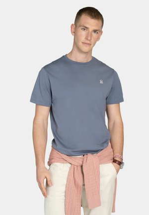 Joven con camiseta azul con un pequeño logo, pantalones beige claro y un suéter rosa atado alrededor de la cintura frente a un fondo liso.