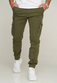 Olive green cargo joggers with tapered cuffs, two side pockets, and zippered accents. Paired with white athletic shoes.