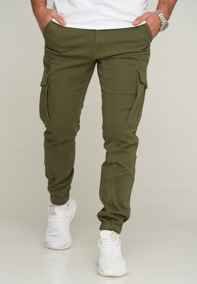 Olive green cargo joggers with tapered cuffs, two side pockets, and zippered accents. Paired with white athletic shoes.