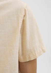 Korte mouw, lichtbeige shirt van textuurstof. Heeft een nette stiksel langs de mouwzoom, met een zachte en ademende uitstraling.