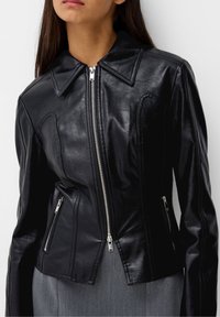 Czarna skórzana kurtka z szerokim kołnierzem, wyraźnym zapięciem na zamek oraz bocznymi kieszeniami z srebrnymi detalami. Dopasowany krój i gładka tekstura.