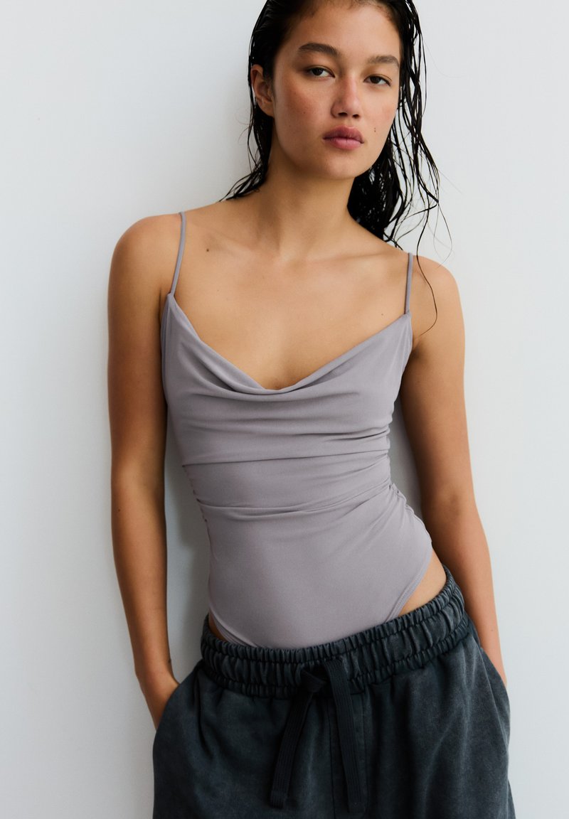 Jeune femme aux cheveux mouillés portant un haut sans manches gris clair et un pantalon gris foncé avec cordon, debout contre un mur blanc.