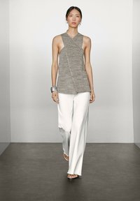 Graues ärmelloses Oberteil mit Wickeldesign und strukturiertem Strickstoff, kombiniert mit lockeren weißen Hosen. Das Model trägt ein metallisches Armreif.