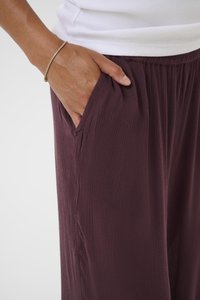 Hand with gold bracelet resting in pocket of textured dark purple pants, wearing white ribbed top.
