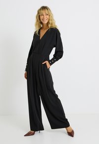 Mono negro con escote en V profundo, mangas largas, diseño plisado, bolsillos laterales y detalles de botones en los puños. Textura de tela suave.