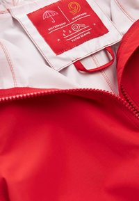 Fermeture éclair d'une veste rouge partiellement ouverte montrant l'étiquette intérieure avec des icônes et du texte indiquant imperméable, coupe-vent et coutures étanches sur tissu blanc.