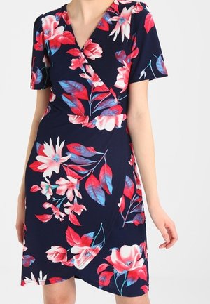 Femme portant une robe portefeuille bleu marine à manches courtes, ornée de grands motifs floraux et feuillus rouges, roses et blancs.