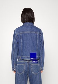 Džínová bunda v středně modré barvě s klasickým límcem, dvěma náprsními kapsami a vybledlou texturou. Na zádech má modrou značkovou cedulku.