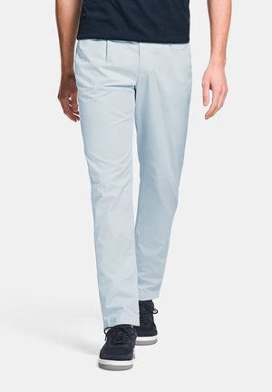 Man draagt lichtblauwe broek en zwarte sneakers, loopt naar voren tegen een witte achtergrond.