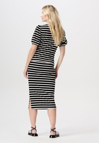 Vrouw met lang blond haar in een zwart-wit gestreepte midi jurk en zwarte platte sandalen, staande tegen een witte achtergrond.